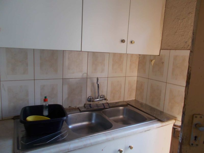 A compact kitchen area featuring a double stainless steel sink, wall-mounted faucet, tiled backsplash, and functional upper and lower cabinetry.