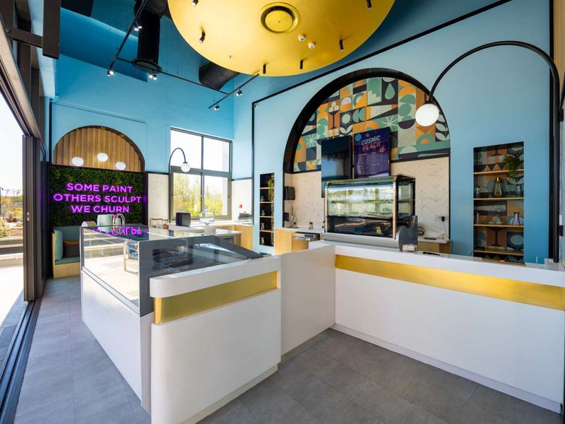 Modern commercial service area featuring a sleek white and gold counter, display refrigeration, vibrant blue walls, and large sliding doors opening to the exterior.