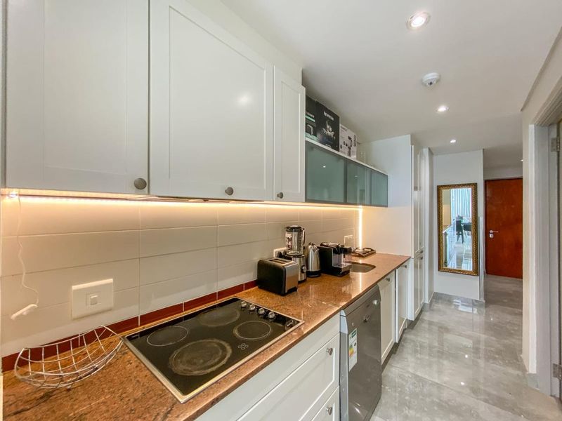 A modern galley-style kitchen featuring white cabinetry, stone countertops, an integrated electric hob, and sleek tiled backsplash with ambient under-cabinet lighting.