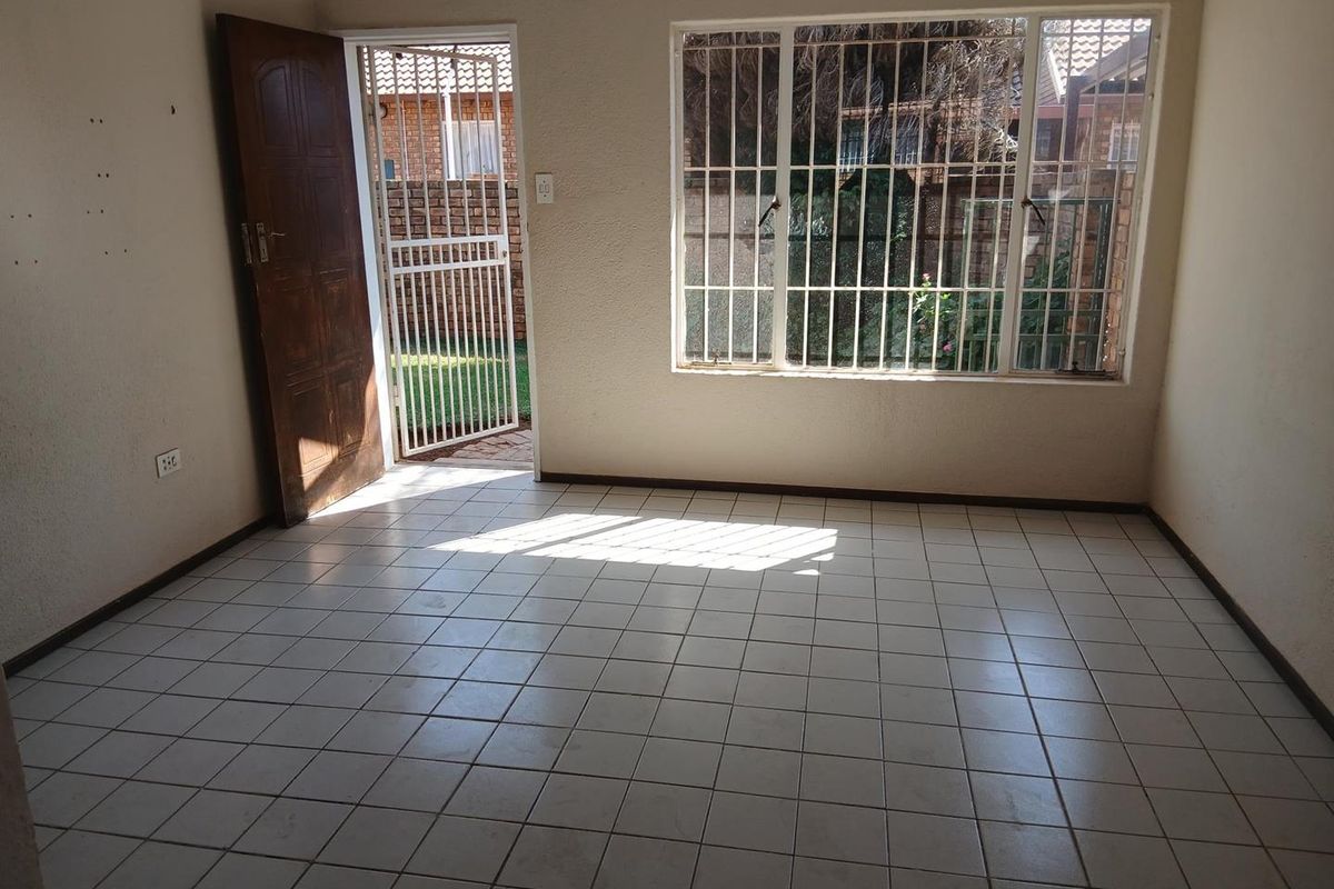 An empty living room featuring tiled flooring, a timber door leading to the exterior, and large windows fitted with security burglar bars.