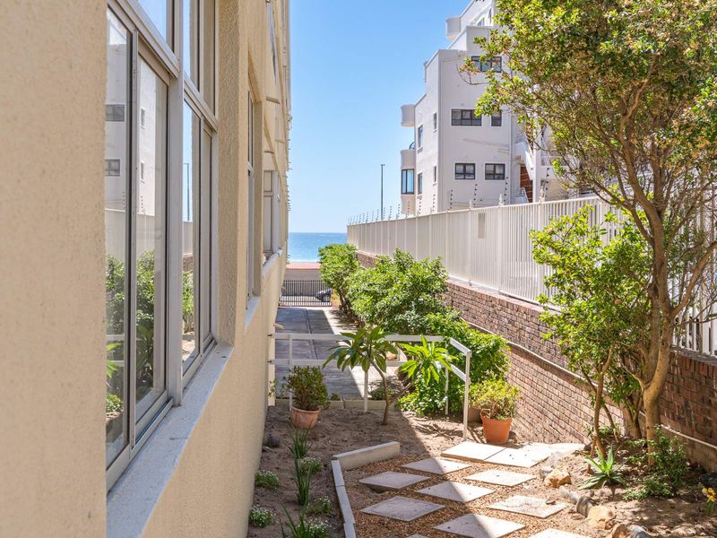 A side exterior view of the apartment building featuring a paved walkway, brick boundary wall, and security fencing with a glimpse of the ocean.