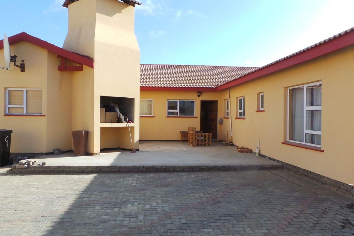 Rear exterior view featuring a paved courtyard, built-in braai area, and tiled roof structure with yellow painted walls.