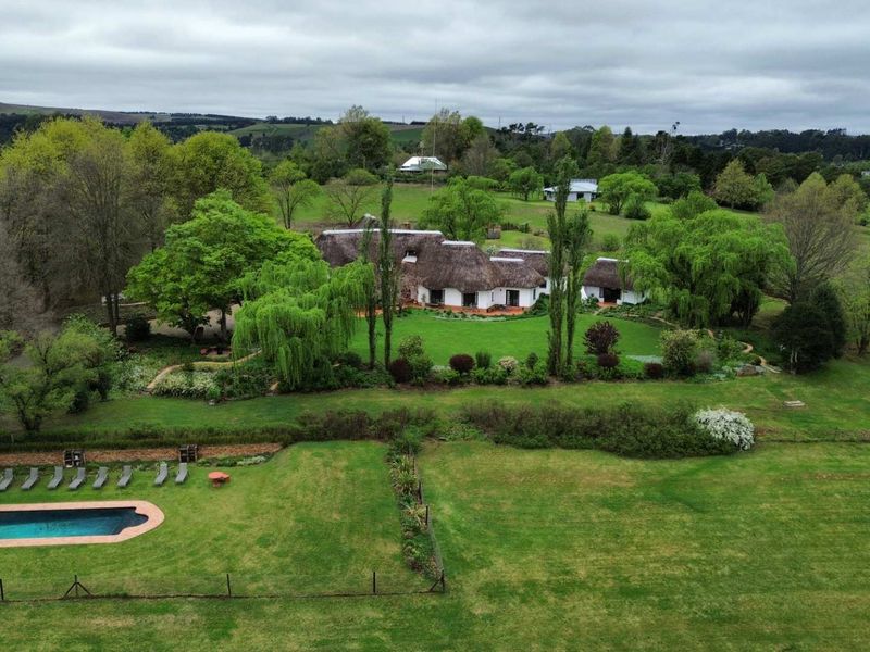 Aerial view of a rural property featuring a traditional thatched-roof house, expansive landscaped gardens, and a private swimming pool set in a scenic landscape.