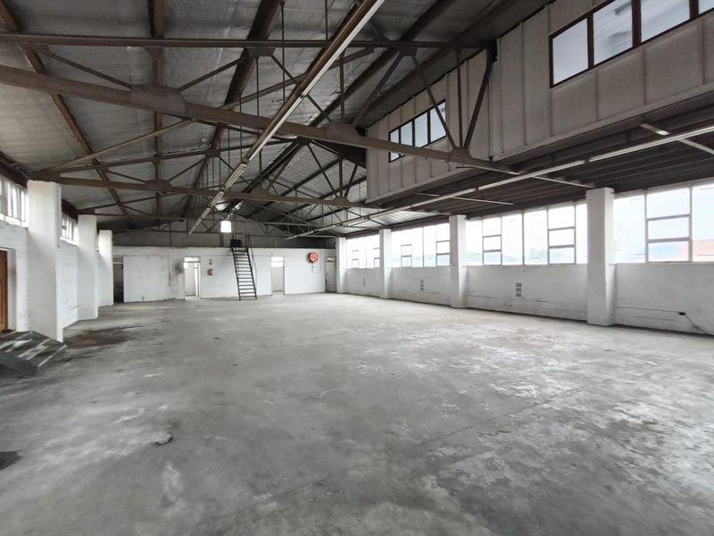 Spacious industrial warehouse area featuring high exposed steel roof trusses, concrete flooring, and large windows providing ample natural light.