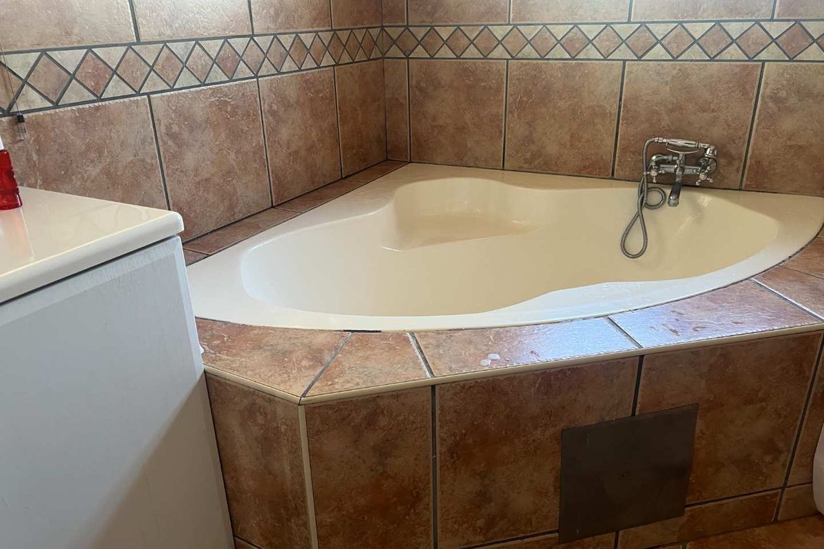 A corner bathtub featuring brown ceramic wall and deck tiles with a decorative border and chrome faucet fixtures.