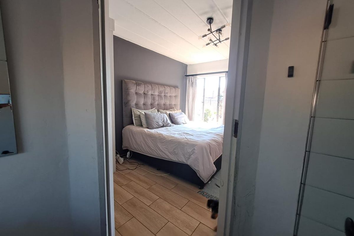 A well-lit bedroom featuring wood-look tile flooring, a dark accent wall, and a modern ceiling light fixture, viewed through a doorway.