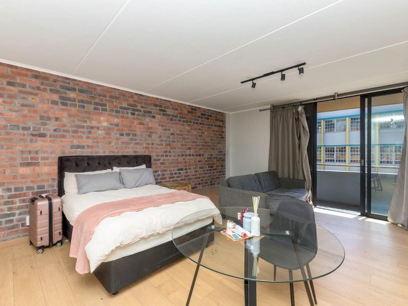 Spacious bedroom featuring an exposed brick feature wall, light laminate flooring, and direct access to a private balcony through sliding glass doors.