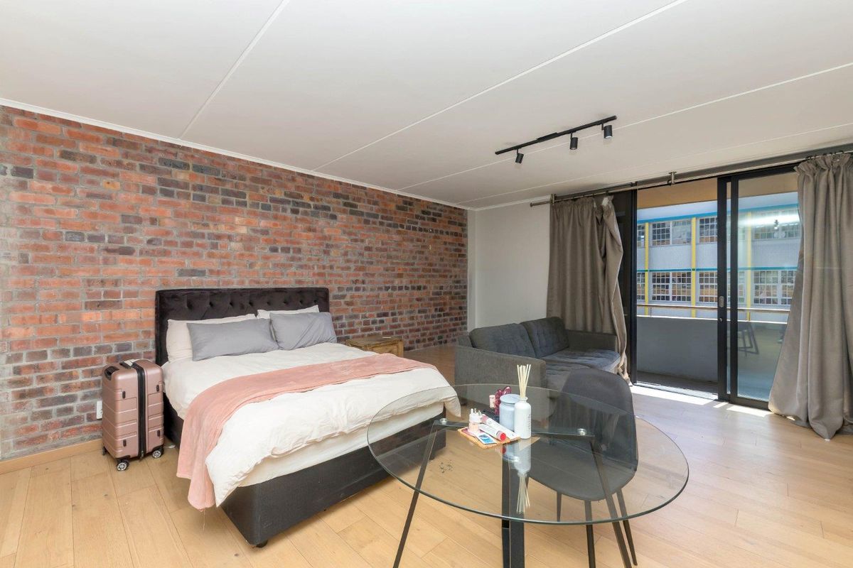 Spacious bedroom featuring an exposed brick feature wall, light laminate flooring, and direct access to a private balcony through sliding glass doors.