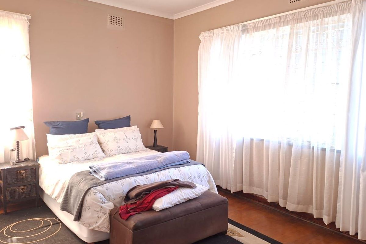 A well-lit bedroom featuring polished timber flooring, neutral wall tones, and large windows with sheer curtains, providing a comfortable and functional living space.