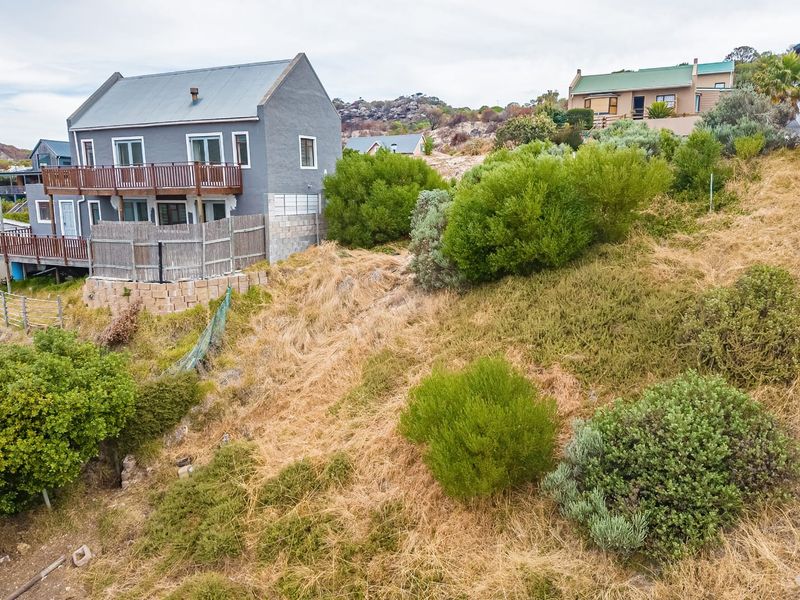An elevated view of a sloped, undeveloped residential plot featuring natural vegetation and a retaining wall, adjacent to existing residential properties.