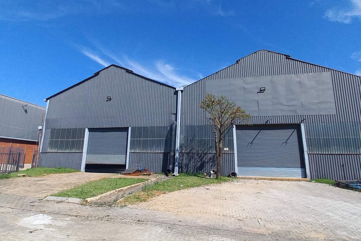 Industrial warehouse facility featuring dual roller shutter doors, corrugated metal exterior cladding, and a paved forecourt area.