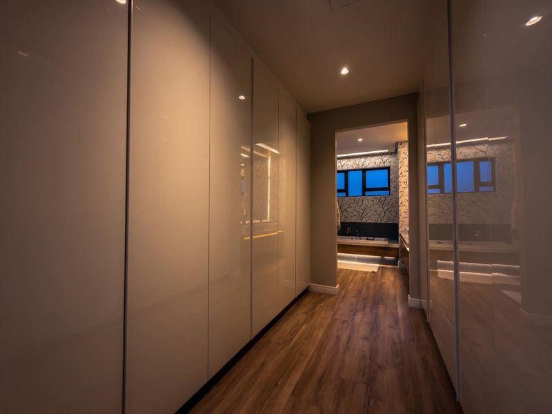 A modern hallway featuring high-gloss built-in cupboards, warm laminate flooring, and recessed downlighting, leading into a bathroom area.