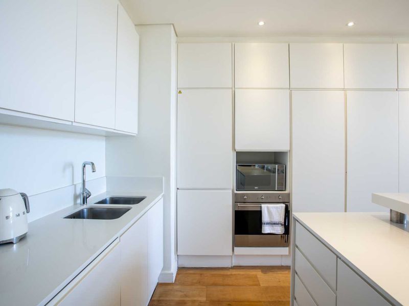 Modern kitchen featuring sleek white cabinetry, stone countertops, integrated appliances, double sink, and warm hardwood flooring with recessed lighting.