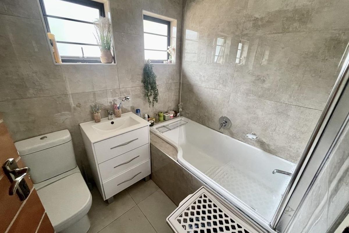 A modern bathroom featuring neutral floor-to-ceiling tiling, a built-in vanity with basin, a toilet, and a bathtub with an integrated shower screen.