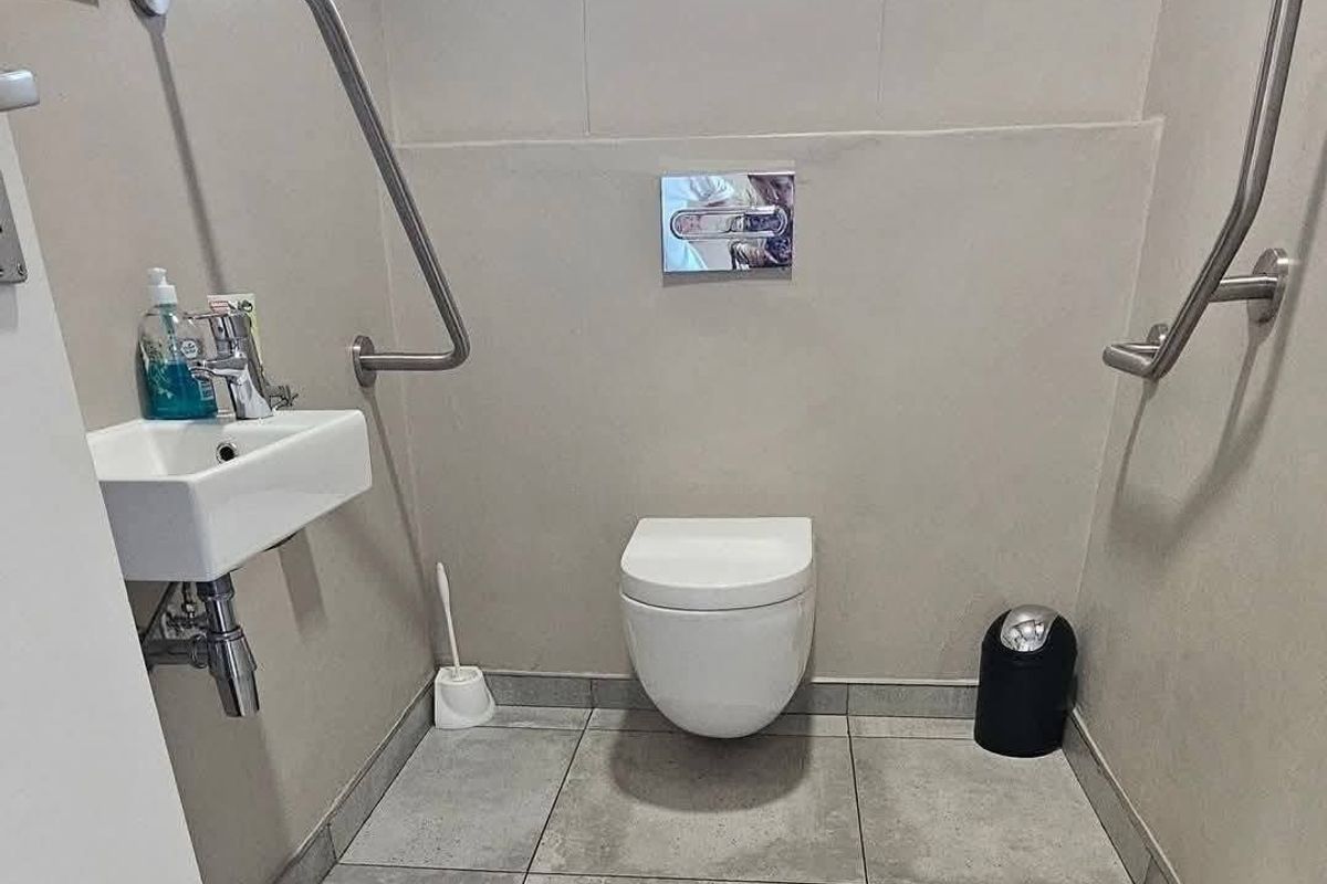 A functional, accessible restroom featuring a wall-mounted toilet, a small wall-hung basin, and stainless steel grab rails for accessibility.