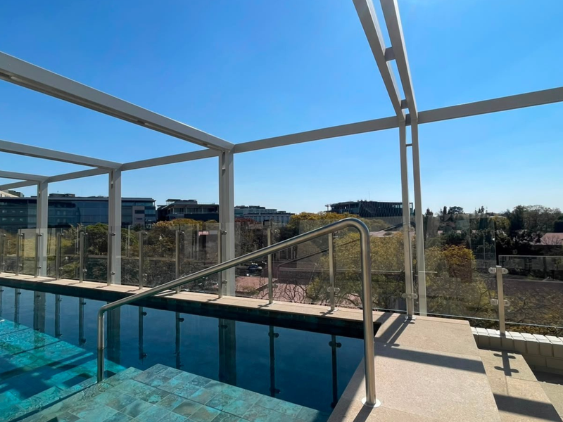 A modern rooftop swimming pool featuring a glass balustrade, metal pergola structure, and clear views of the surrounding area.