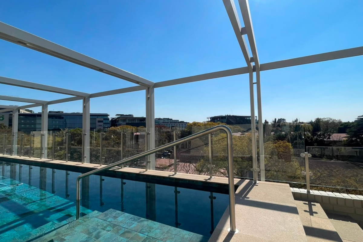 A modern rooftop swimming pool featuring a glass balustrade, metal pergola structure, and clear views of the surrounding area.