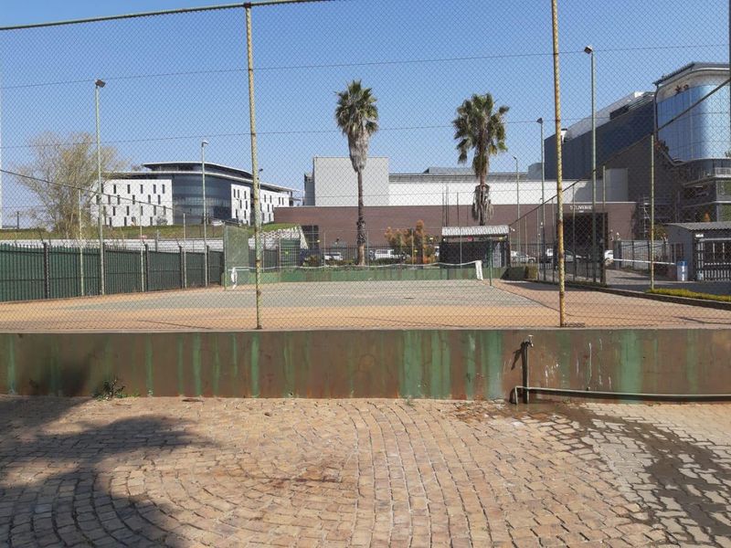 A communal tennis court facility featuring a fenced playing area, floodlights, and paved surroundings within the apartment complex.