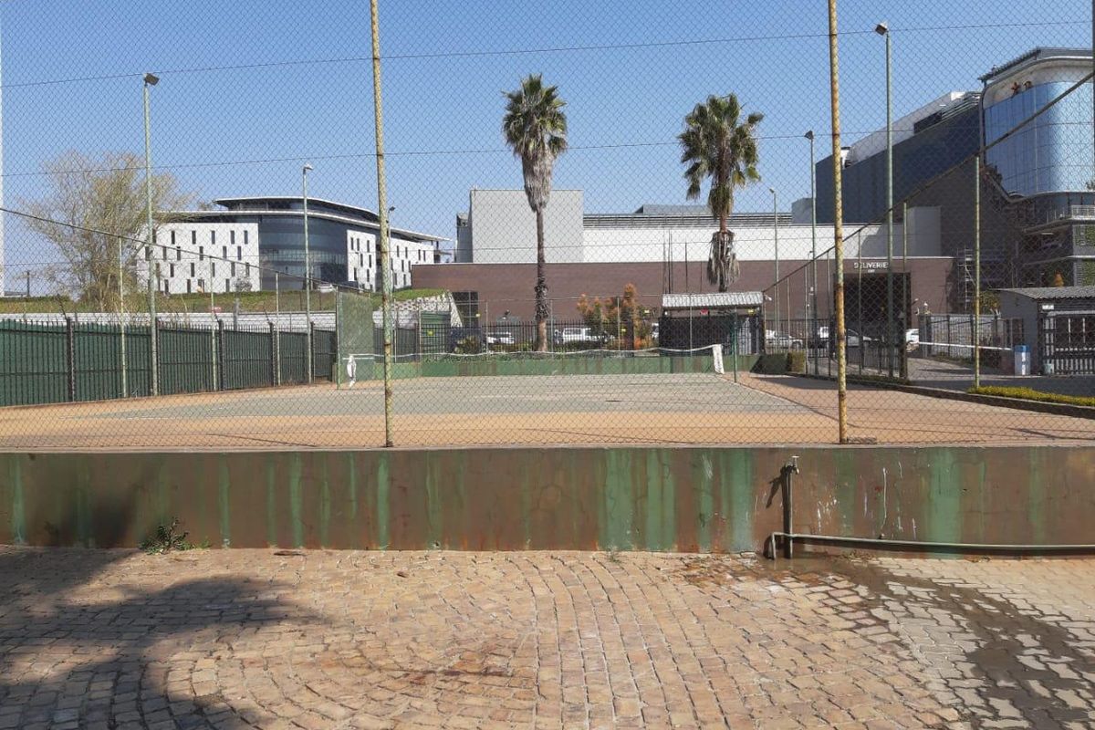 A communal tennis court facility featuring a fenced playing area, floodlights, and paved surroundings within the apartment complex.