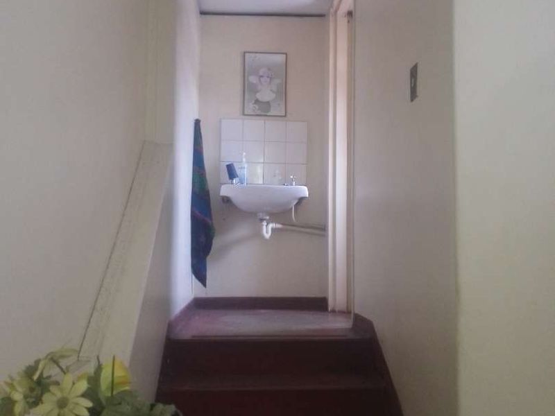 A narrow hallway featuring a small wall-mounted basin with a tiled splashback, leading to a staircase with worn flooring.