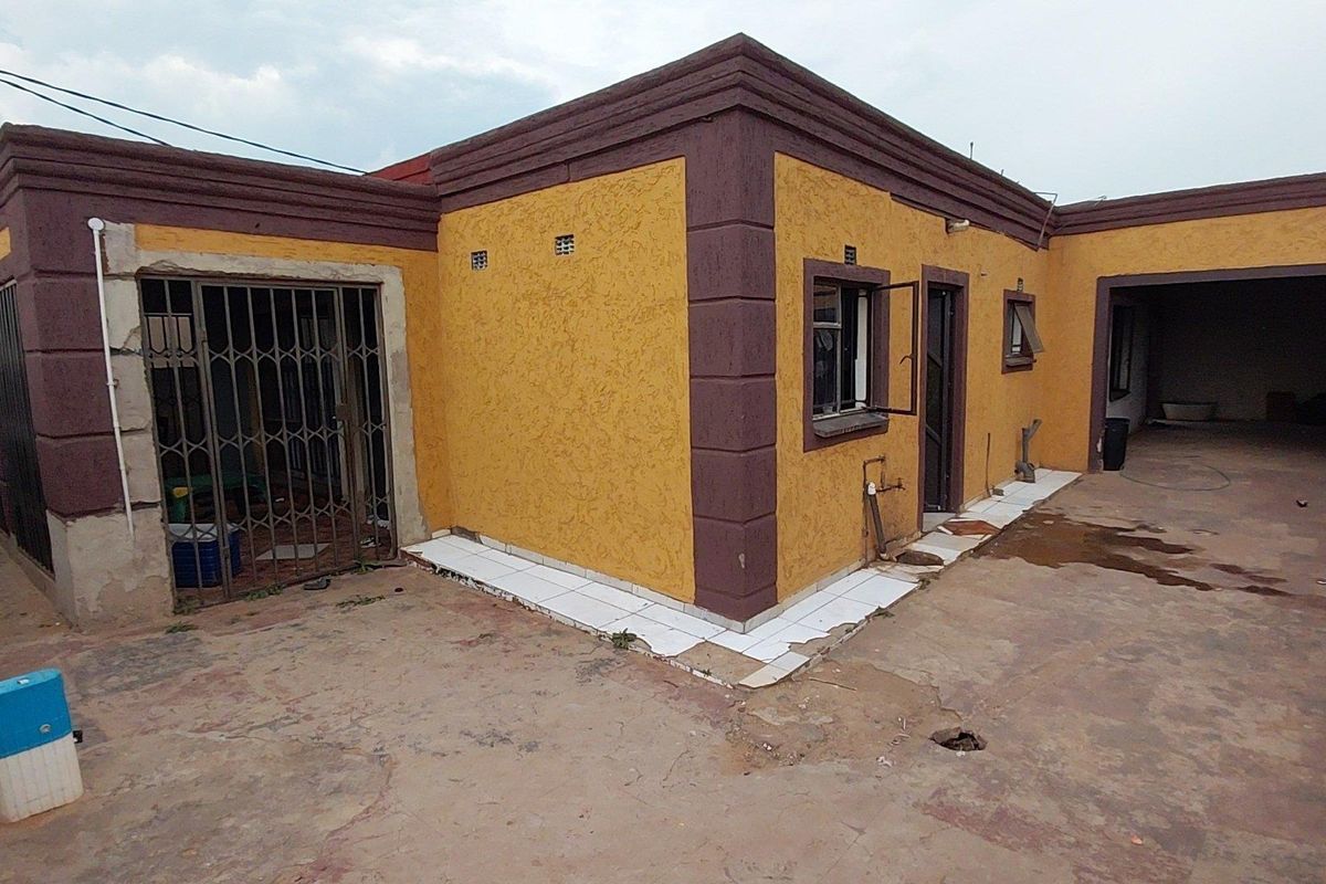 Rear exterior view of the house featuring a textured facade, security gate, and a covered patio area with worn concrete and damaged tiling.