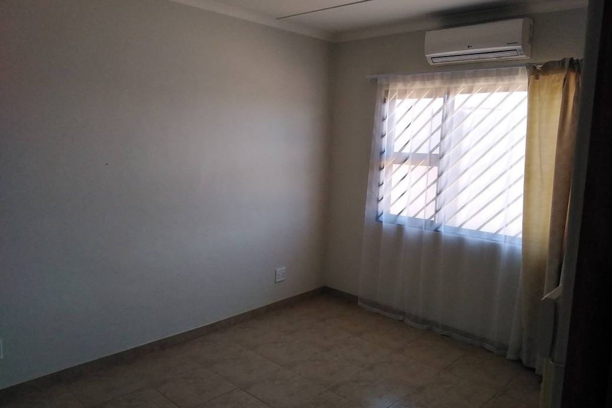 A clean, empty bedroom featuring tiled flooring, a wall-mounted air conditioning unit, and a window fitted with security bars.