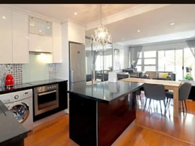 Modern open-plan kitchen and living area featuring sleek cabinetry, stone countertops, integrated appliances, and hardwood flooring with access to a balcony.