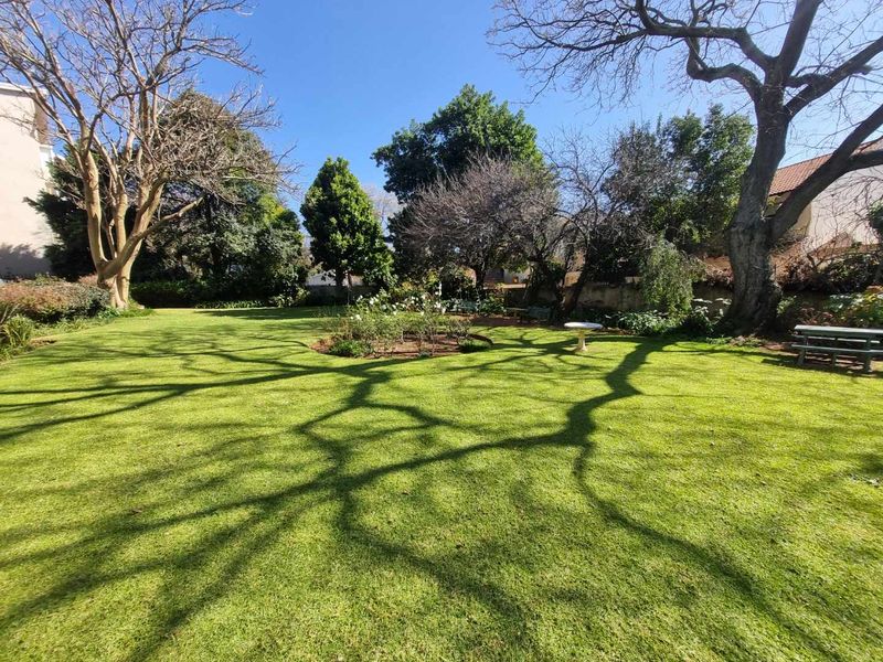 A spacious, well-maintained communal garden featuring mature trees, expansive lawn areas, and established flower beds within a secure apartment complex setting.