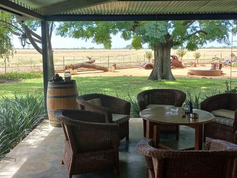 A covered outdoor patio area featuring concrete flooring and metal roofing, overlooking a spacious farm garden and open landscape.
