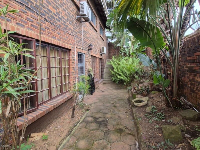 A side exterior view of the property featuring a brick facade, paved walkway, and established garden area with boundary walling.