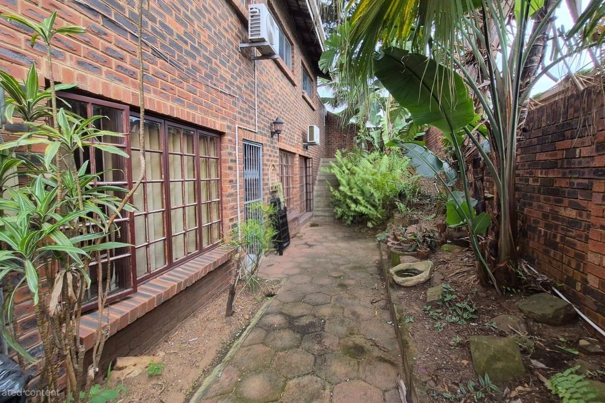 A side exterior view of the property featuring a brick facade, paved walkway, and established garden area with boundary walling.