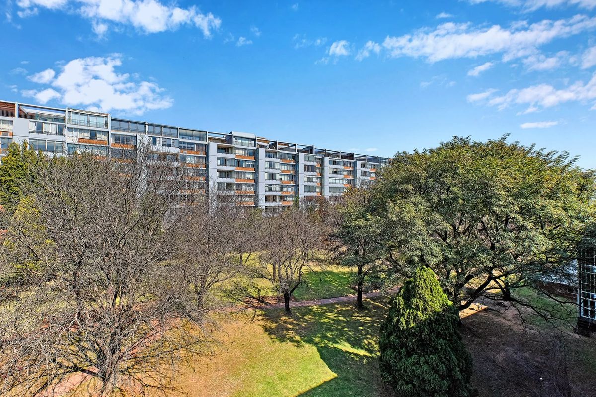 A view of the rear elevation of a multi-story apartment complex overlooking a well-maintained garden with mature trees and paved walkways.