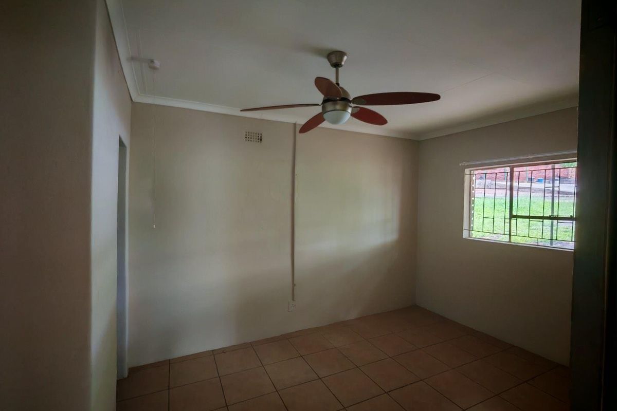 A simple, empty bedroom featuring tiled flooring, a ceiling fan, and a window with security bars.