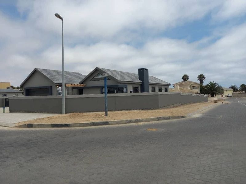 Street view of a modern single-story house featuring a grey exterior, tiled roof, chimney, and a perimeter boundary wall with a paved driveway.