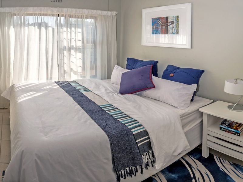 A well-lit bedroom featuring neutral wall tones, tiled flooring, and large windows with sheer curtains, providing a bright and comfortable living space.
