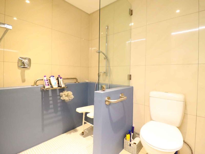 A functional bathroom featuring a walk-in shower with a glass screen, grab rails for accessibility, and neutral wall tiling.