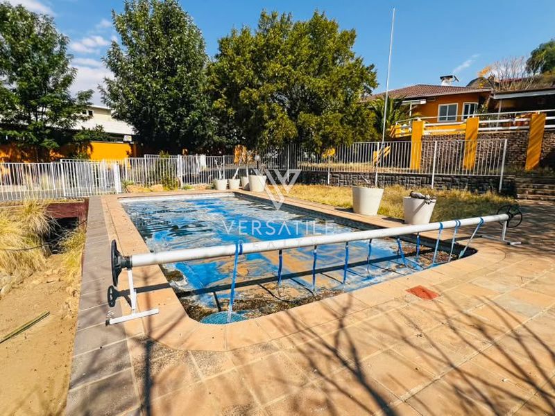 Outdoor pool area featuring a paved surround, metal perimeter fencing, and a pool cover, set against a terraced garden backdrop.
