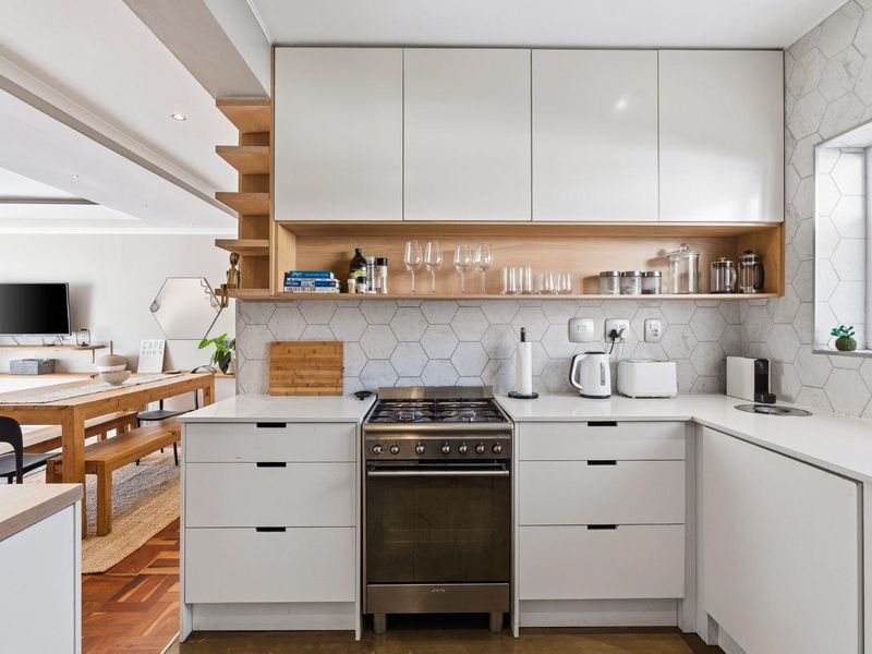Modern open-plan kitchen featuring sleek white cabinetry, stone countertops, a gas stove, and a stylish hexagon tile backsplash, seamlessly flowing into the dining area.