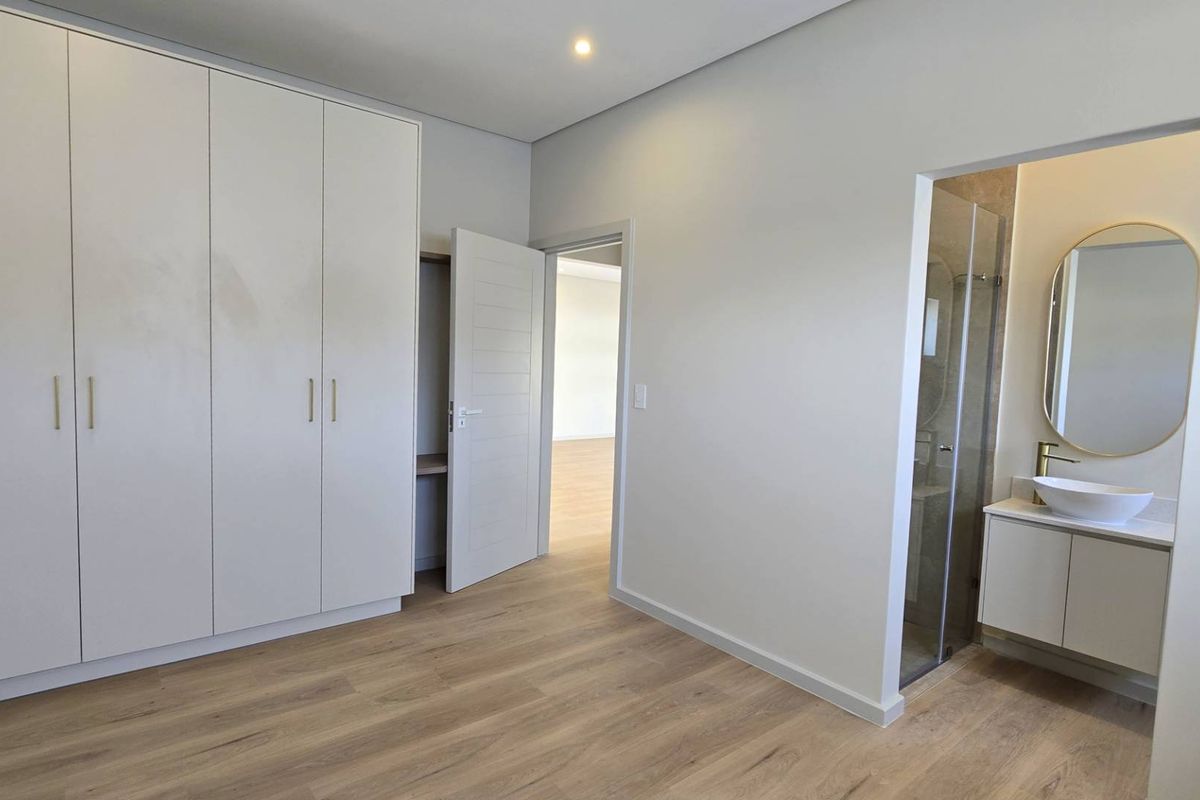Modern bedroom featuring light laminate flooring, ample built-in cupboard space, and a sleek en-suite bathroom with a vanity basin and shower.