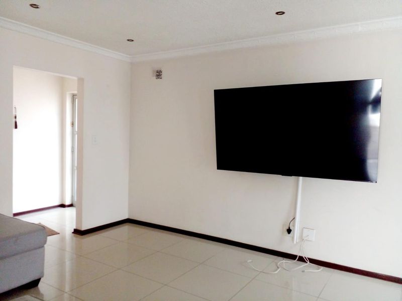 A bright living room featuring neutral tiled flooring, recessed downlighting, and coving, with a wall-mounted television and visible cable management.