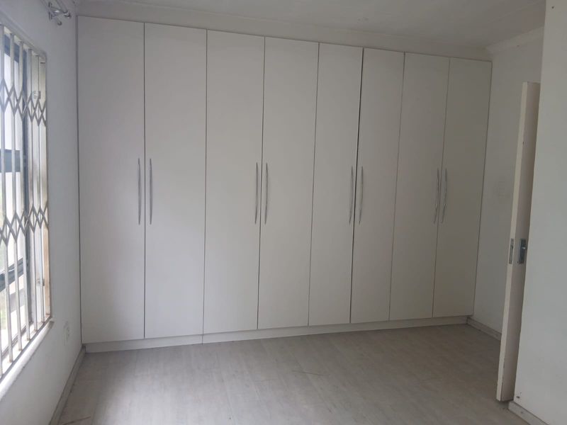 A clean, empty bedroom featuring extensive white built-in cupboards, light-toned laminate flooring, and a window fitted with a security gate.