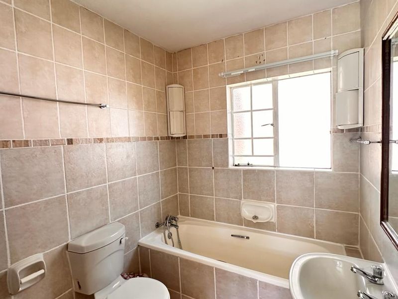 A functional bathroom featuring a bathtub, pedestal basin, and toilet, finished with neutral wall tiling and a window for natural light.