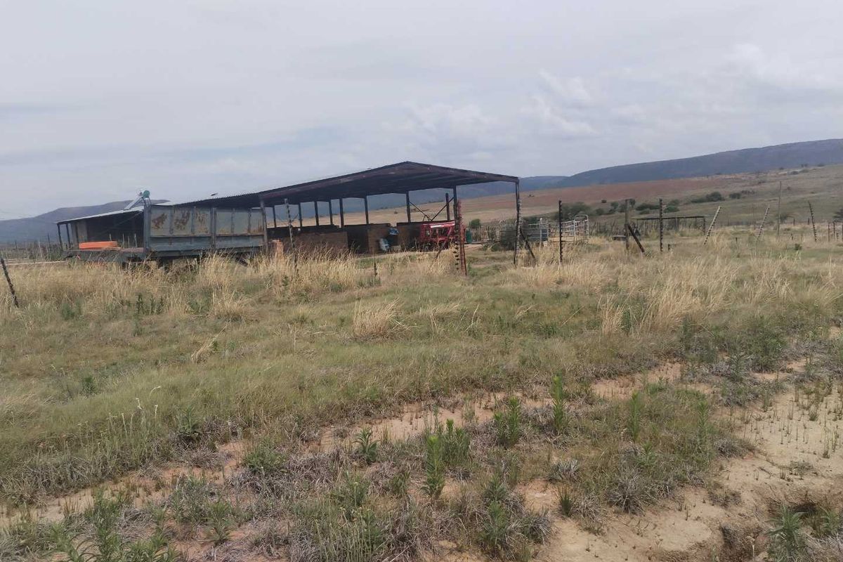 An open-sided farm shed structure situated on a large, undeveloped rural plot with surrounding fencing and distant mountain views.