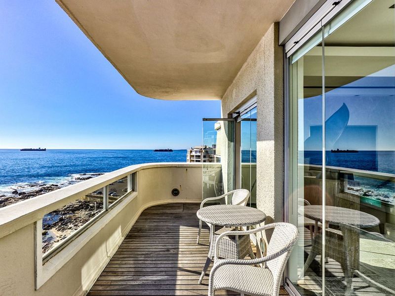 A spacious balcony featuring timber decking, a glass balustrade, and sliding glass doors, offering panoramic ocean views.