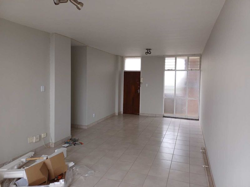 An empty, open-plan living area featuring neutral tiled flooring, a secure entrance door, and large glass panels providing natural light.
