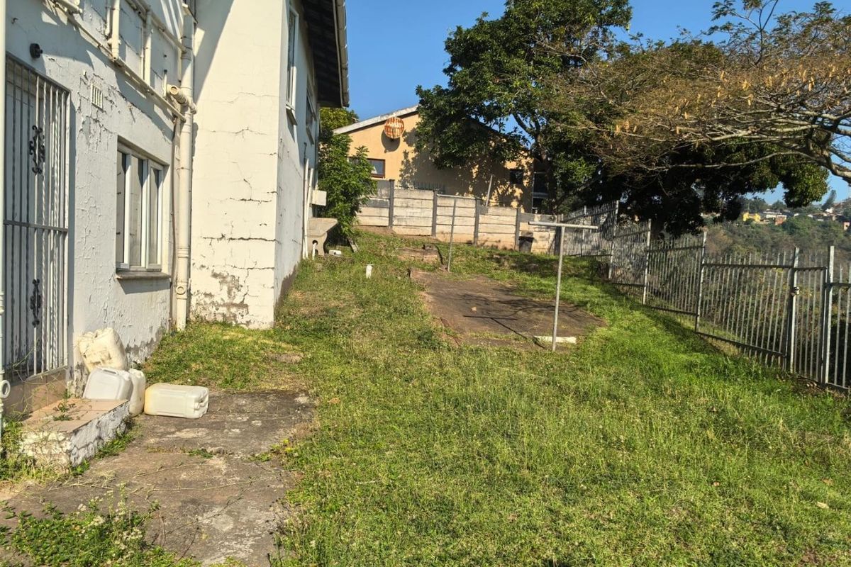 Rear exterior view of the property showing a grassed yard, concrete paving, and perimeter fencing with visible wear to the building's exterior wall finish.