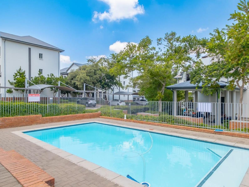 A well-maintained communal swimming pool area featuring brick paving, metal fencing, and a covered gazebo within an apartment complex.