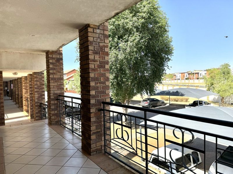 A spacious covered balcony featuring brick pillars, tiled flooring, and a decorative metal balustrade overlooking the complex parking area.