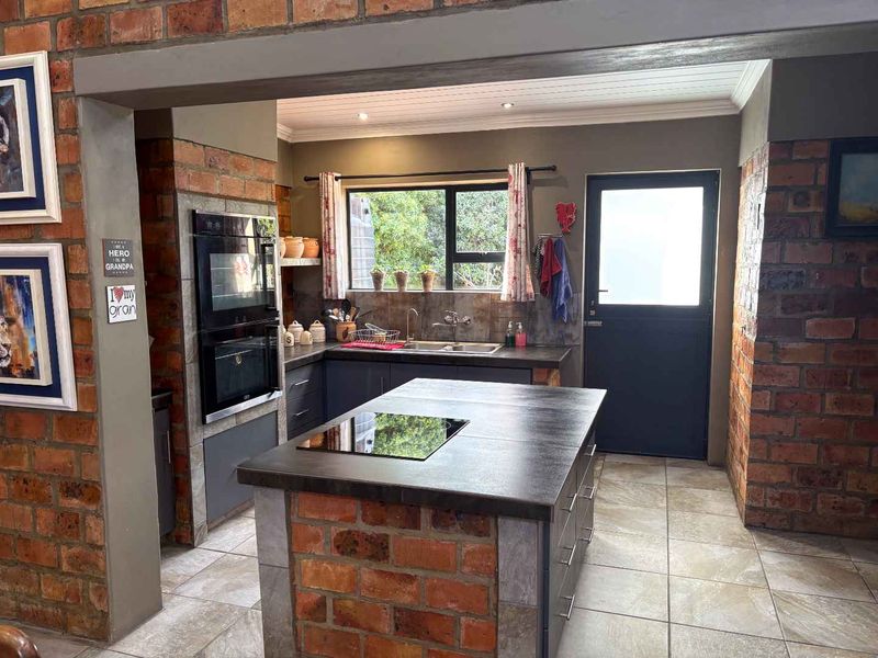 A rustic kitchen featuring exposed brick pillars, a central island with a built-in hob, dark cabinetry, and a double wall oven.