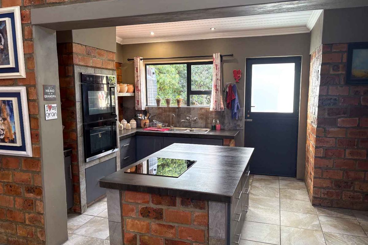 A rustic kitchen featuring exposed brick pillars, a central island with a built-in hob, dark cabinetry, and a double wall oven.